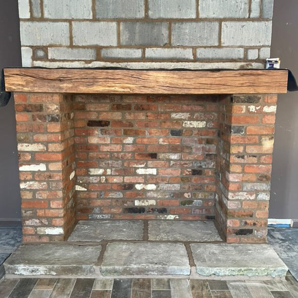 A brick fireplace with a wooden mantel on top, positioned against a wall with visible bricklaying techniques.