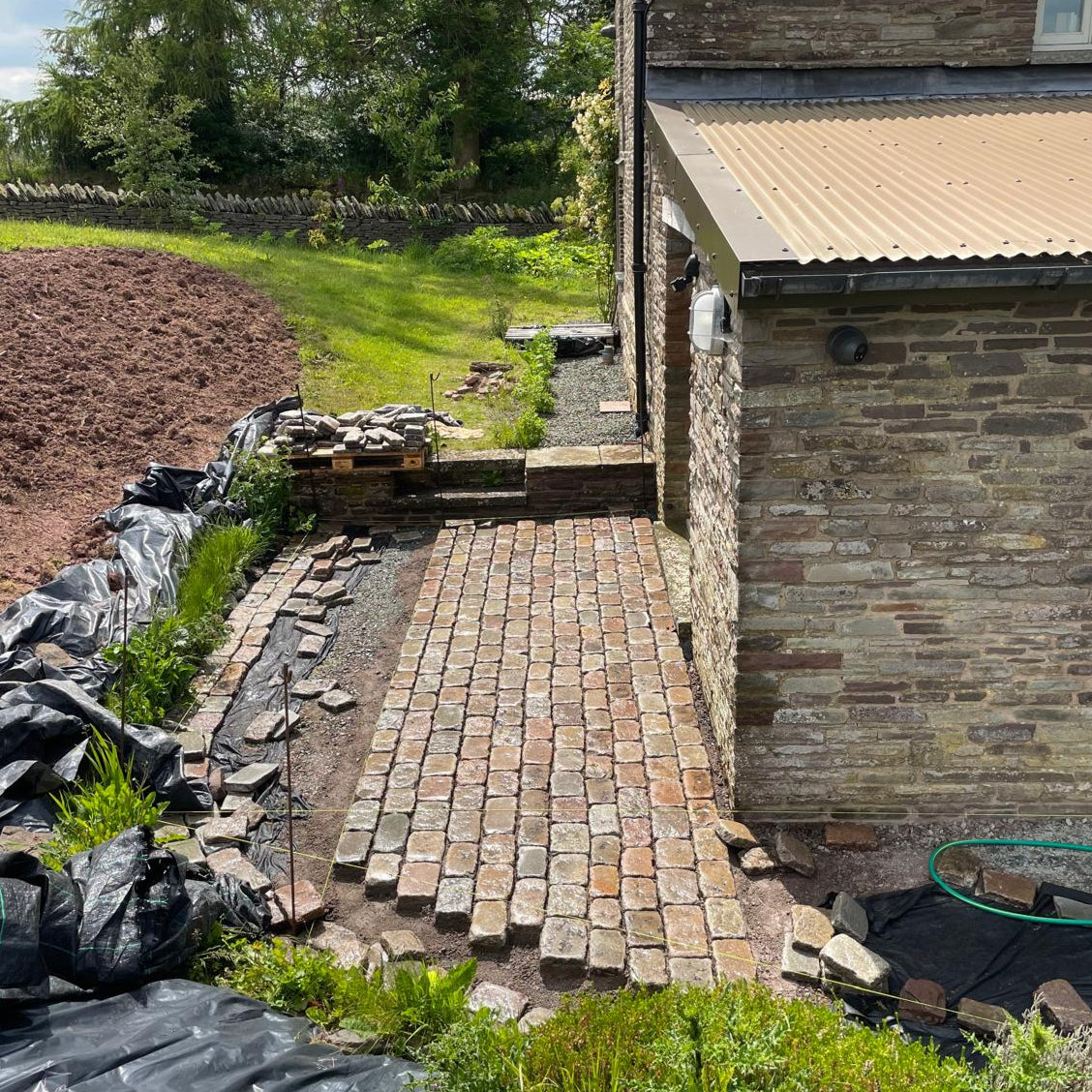 Paver patio construction in a garden area next to a stone building.