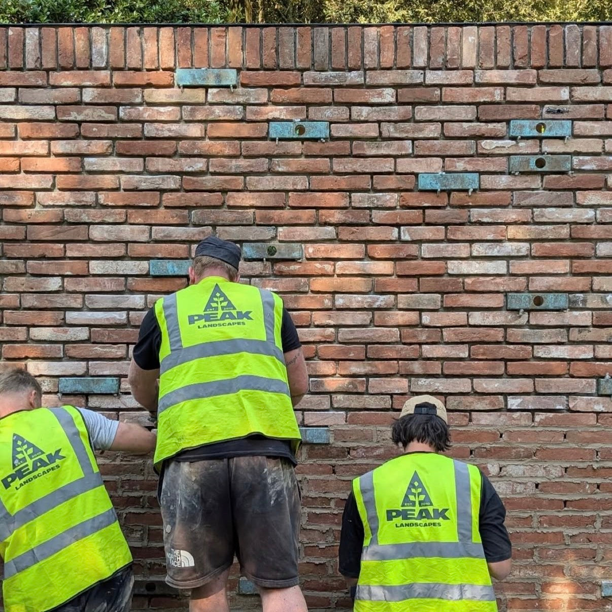 Three workers in Peak Landscapes vests working on a brick wall.